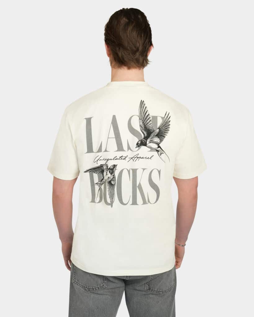 Model wearing the Uncaged Birds Tee in Off-White, back view with large “LAST BUCKS” graphic and flying birds artwork