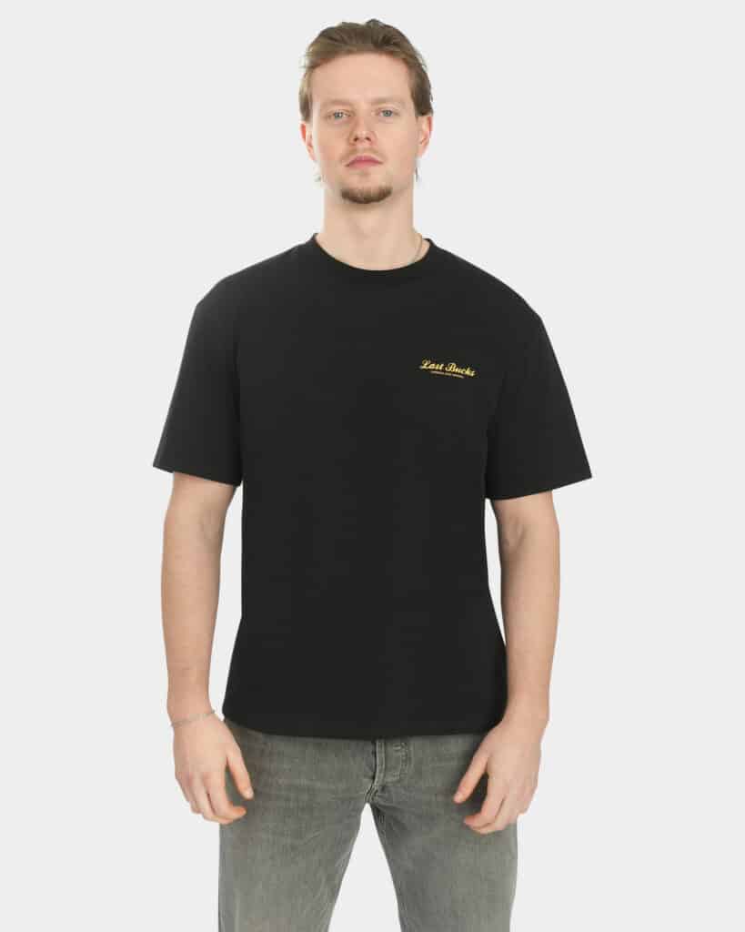 Male model wearing the Script Tee in Jet Black, front view, with a small gold “Last Bucks” chest logo on the left side