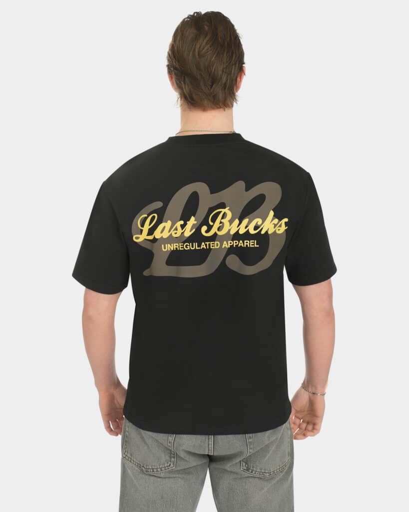 Male model wearing the Script Tee in Jet Black, back view, featuring a large gold “Last Bucks” script print with “Unregulated Apparel” and a big “LB” graphic