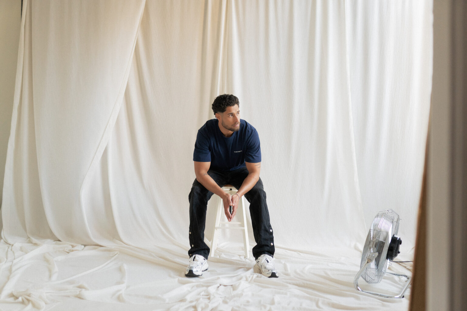 Model wearing the Last Bucks Capital Tee Navy in a studio setting, sitting on a stool.