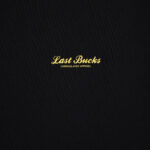 Close-up of the Script Tee in Jet Black featuring a small gold “Last Bucks” script logo with “UNREGULATED APPAREL” underneath on the chest