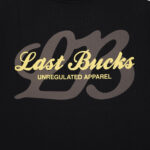 Close-up of the back print on the Script Tee in Jet Black, showing a large tonal “LB” graphic with gold “Last Bucks” script and “UNREGULATED APPAREL” across it
