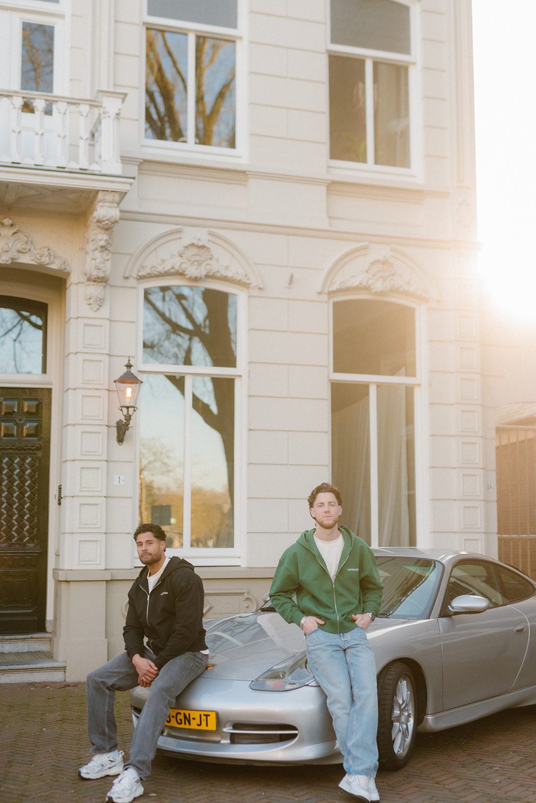 Twee mannen in zip hoodies bij een auto in tegenlicht, de straat die Prada in Amsterdam deelt met andere huizen