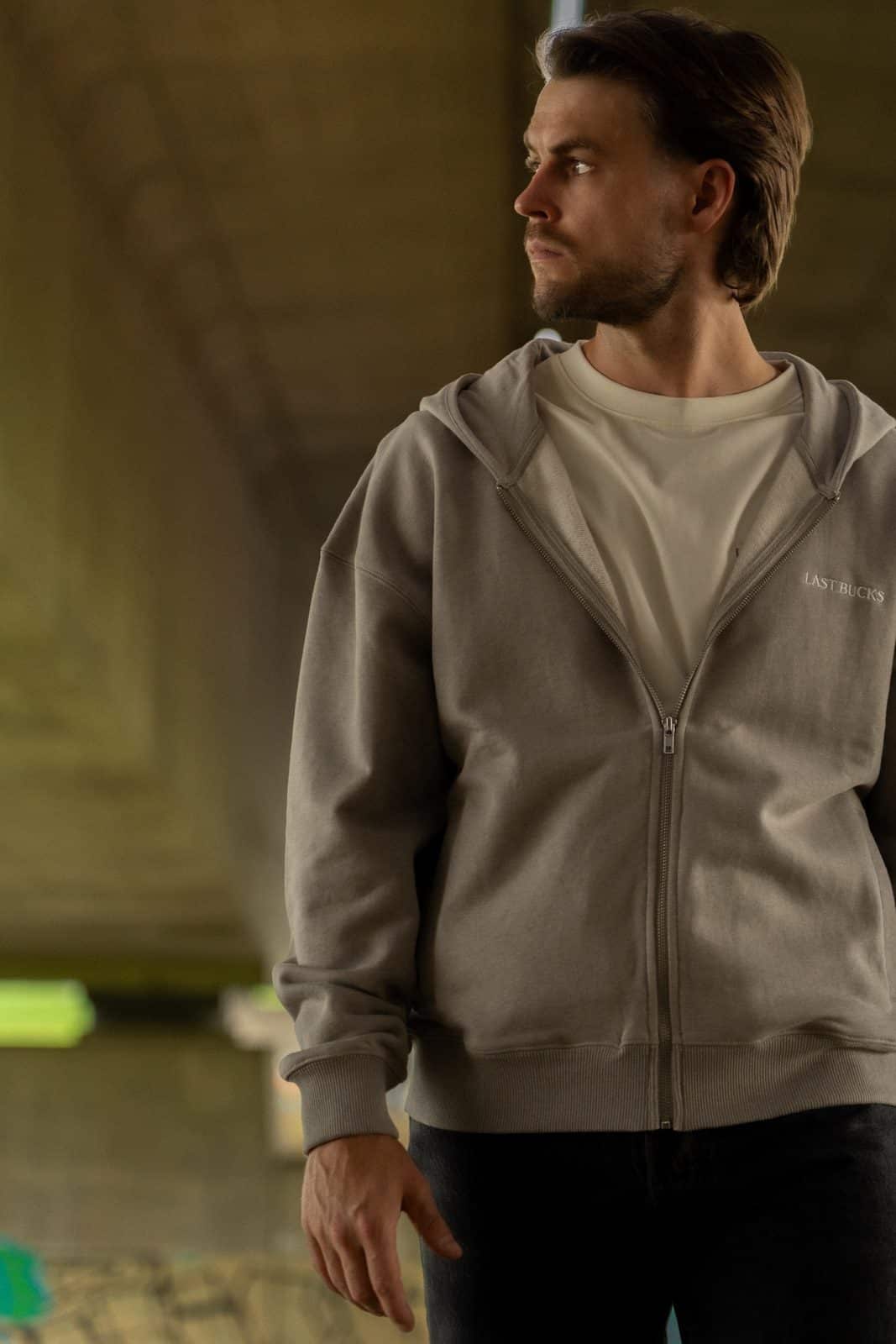 Man in een grijze zip hoodie, half dichtgeritst, over een licht t-shirt en een donkere jeans in een onderdoorgang