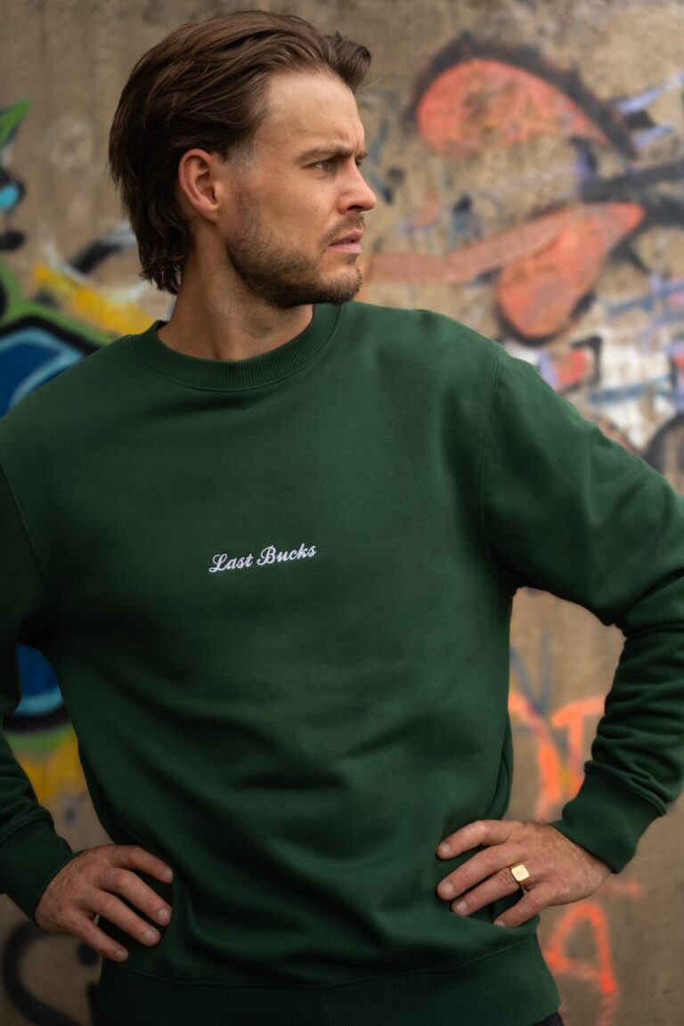 Herfst outfit heren: man in een hunter green crewneck sweater met klein wit script op de borst, handen in de zij, voor een graffitimuur