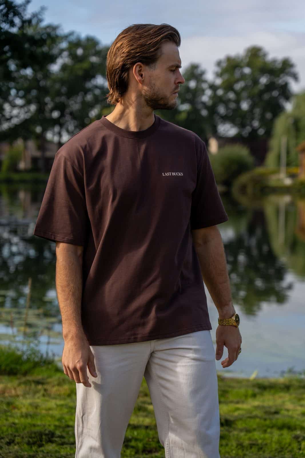 Man in een mocha brown t-shirt met klein woordmerk en een crèmekleurige broek aan de oever van een vijver