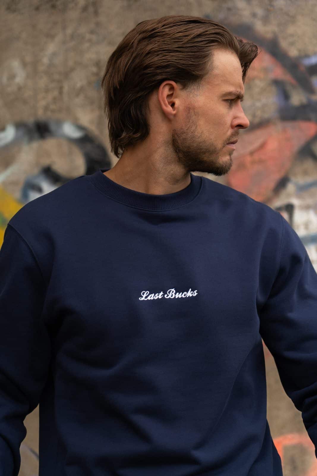 Close-up van een man in een navy crewneck sweater met klein wit script en een geribde boord rond de hals voor een graffitimuur