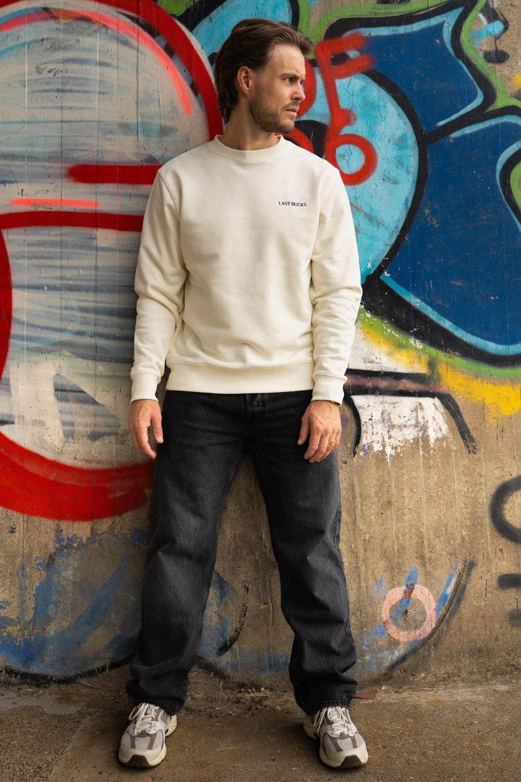 Man in een offwhite crewneck sweater met klein woordmerk, een donkere jeans en sneakers tegen een graffitimuur