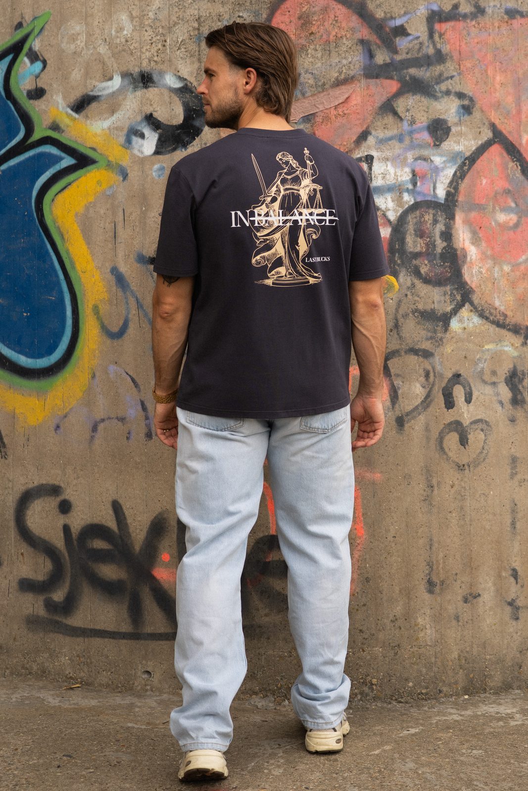 Man van achteren in een donker t-shirt met een goudkleurige rugprint van Vrouwe Justitia en een lichte jeans voor een graffitimuur