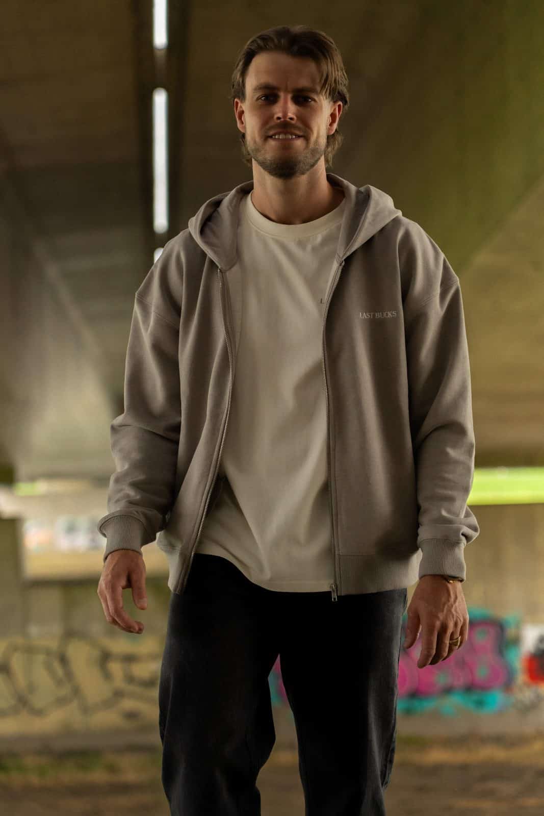 Man in een open grijze zip hoodie over een licht t-shirt dat onder de zoom uitkomt, met een donkere jeans in een onderdoorgang