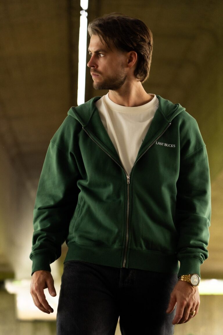Man draagt laagjes: een hunter green zip hoodie, half dichtgeritst, over een offwhite t-shirt in een onderdoorgang met tl-licht