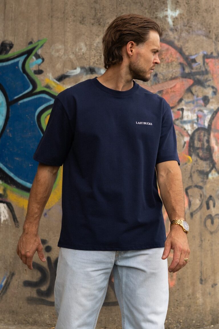 Man in een navy t-shirt met klein woordmerk en een lichte jeans voor een graffitimuur, bij de maattabel heren van Last Bucks