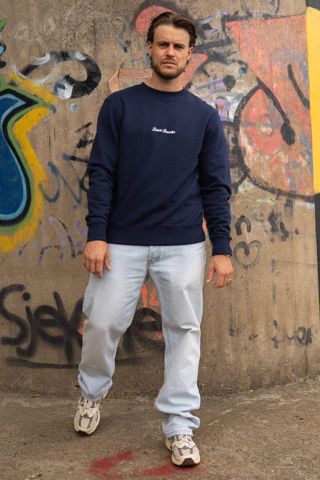 Man in een navy crewneck sweater met klein script, een lichte jeans en witte sneakers voor een graffitimuur
