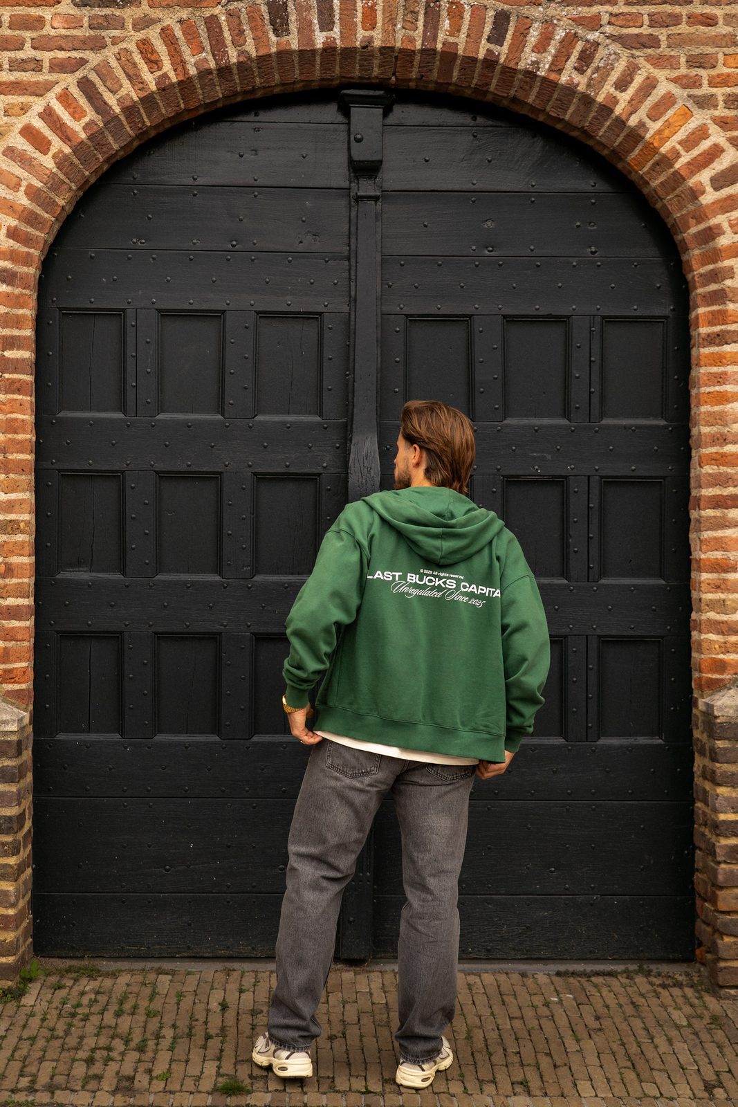 Man van achteren in een hunter green zip hoodie met rugprint en een grijze jeans voor een zwarte boogdeur in een bakstenen muur