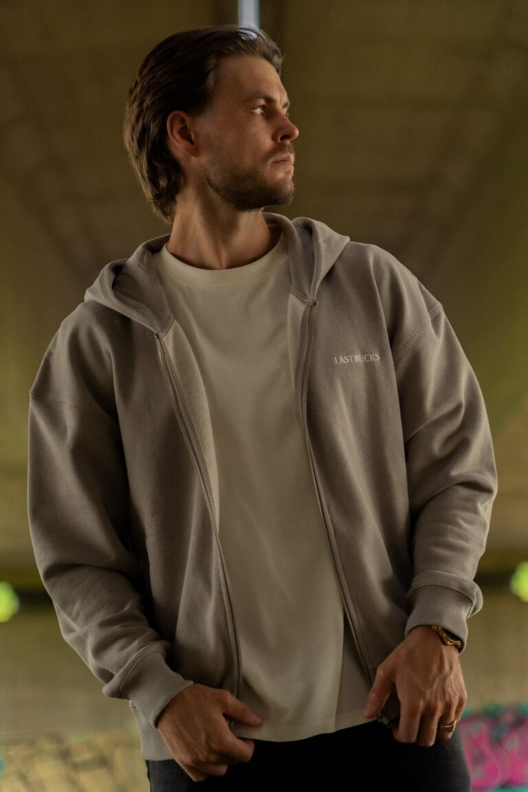 Man in een open grijze zip hoodie met klein woordmerk op de borst over een lichte t-shirt in een onderdoorgang, bij merken zoals Cold Culture