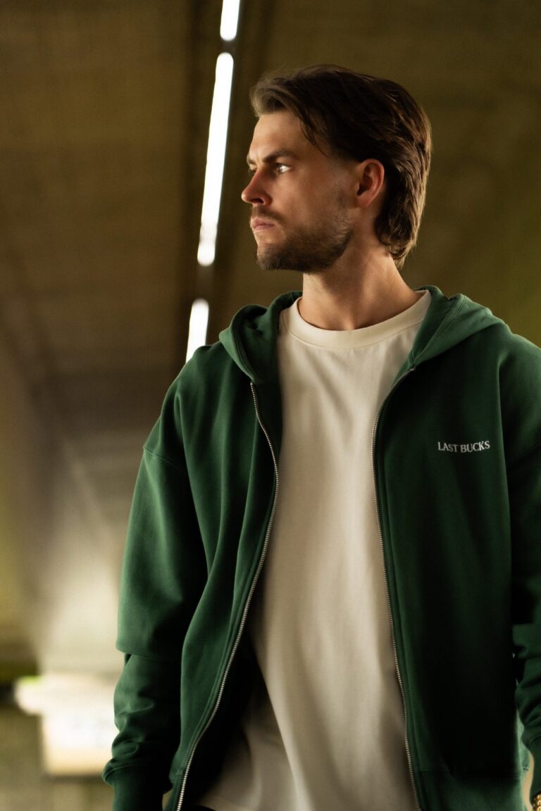 Man in een open donkergroene zip hoodie met klein woordmerk op de borst over een lichte t-shirt in een onderdoorgang, bij merken zoals Scuffers