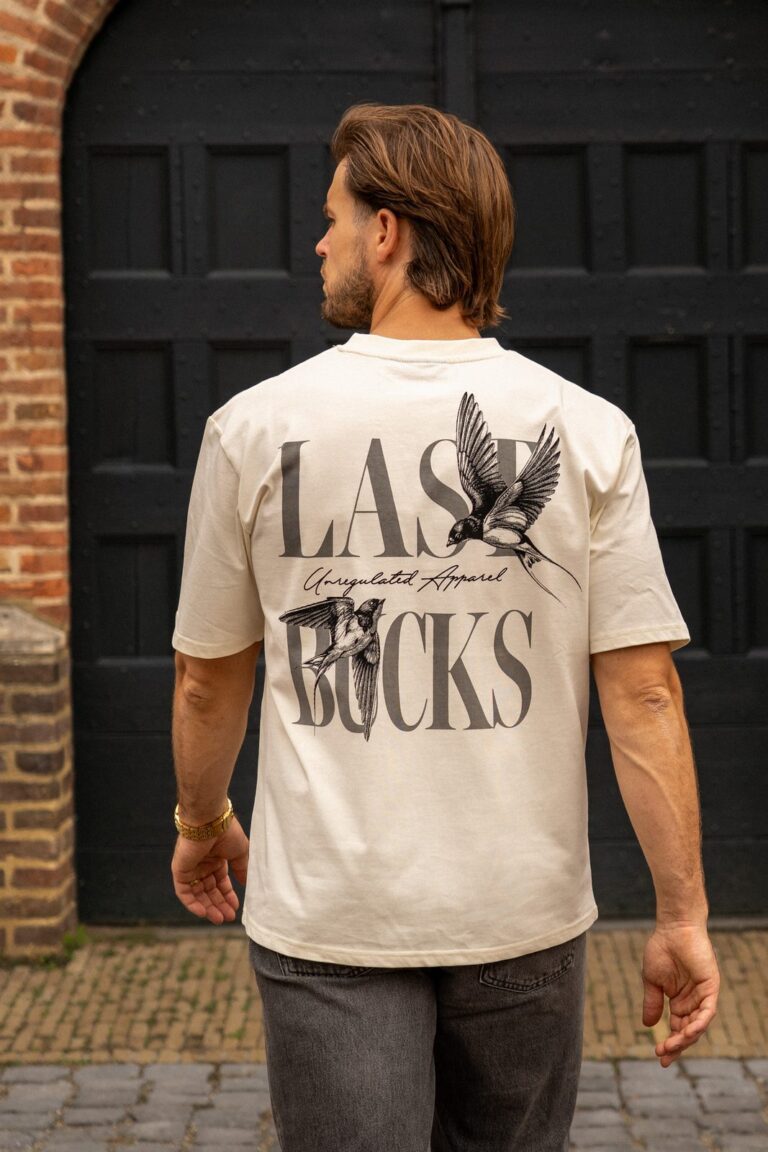 Man op de rug gezien in een offwhite t-shirt met een rugprint van twee vogels en grote letters, voor een zwarte deur in een bakstenen poort, bij merken zoals Suspicious Antwerp
