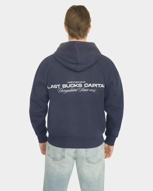 Male model wearing the Last Bucks Capital Zip Hoodie in Navy, shown from the back with the large "LAST BUCKS CAPITAL" back print and "Unregulated Since 2025" script