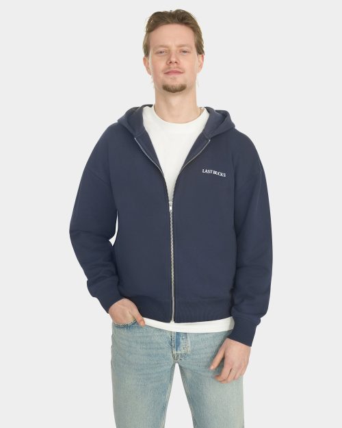Male model wearing the Last Bucks Capital Zip Hoodie in Navy, shown from the front with the zipper partially open and a small "LAST BUCKS" chest logo