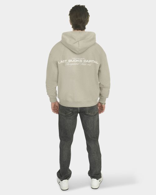 Full body back view of a model wearing The Capital Zip Hoodie in Mari Grey, showing the back print "LAST BUCKS CAPITAL" with "Unregulated Since 2025", paired with grey jeans and white sneakers on a clean background