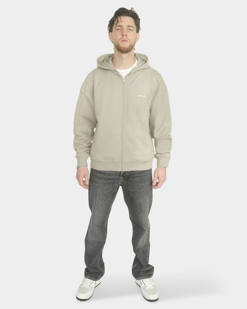 Full body front view of a model wearing The Capital Zip Hoodie in Mari Grey with a small "LAST BUCKS" chest logo, paired with a white t-shirt, grey jeans, and white sneakers on a clean background