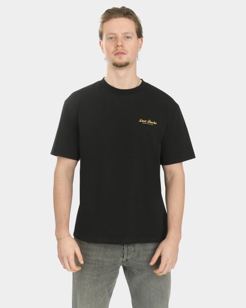 Male model wearing the Script Tee in Jet Black, front view, with a small gold “Last Bucks” chest logo on the left side