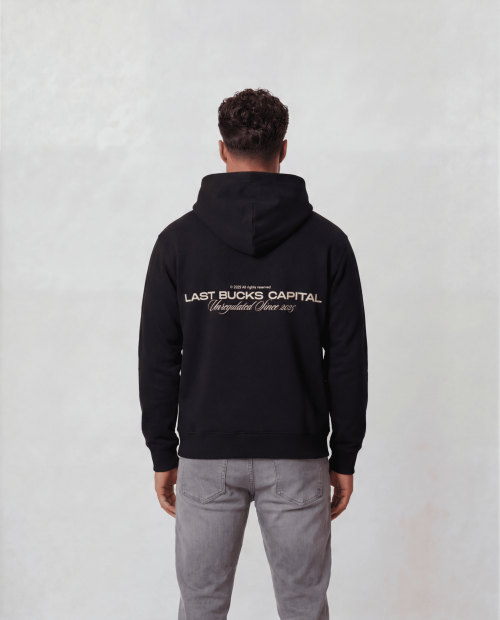 Male model wearing The Capital Zip Hoodie in Jet Black, back view, showing large "LAST BUCKS CAPITAL" print with "Unregulated Since 2025" on the back