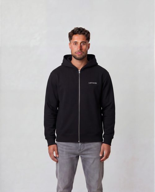 Male model wearing The Capital Zip Hoodie in Jet Black, front view, unzipped over a white t-shirt with small "LAST BUCKS" logo on the chest