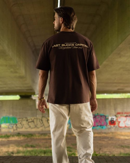The founder wearing the Capital Tee Mocha Brown by Last Bucks, back with the large Last Bucks Capital back print with Unregulated Since 2025, under a concrete overpass with graffiti