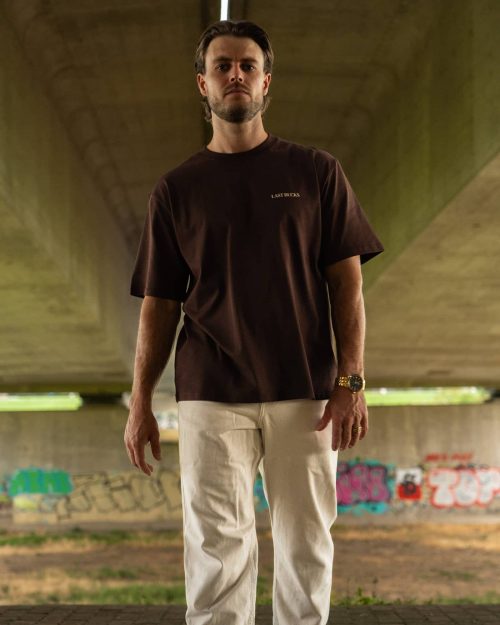 The founder wearing the Capital Tee Mocha Brown by Last Bucks, front with the minimalist chest logo, under a concrete overpass with graffiti