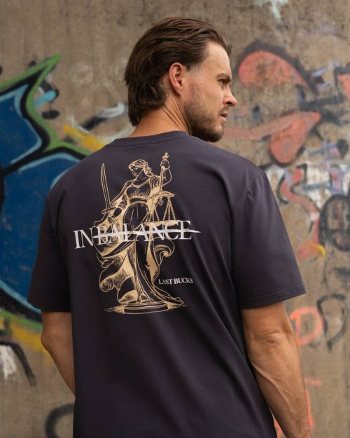 The founder wearing the In Balance Tee Anthracite by Last Bucks, back with the Lady Justice print with her scales and the word balance struck through, against a concrete wall covered in graffiti