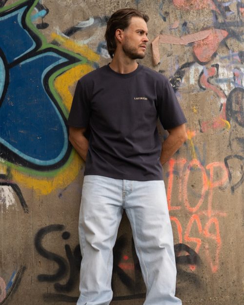The founder wearing the In Balance Tee Anthracite by Last Bucks, front with the minimalist chest logo, against a concrete wall covered in graffiti