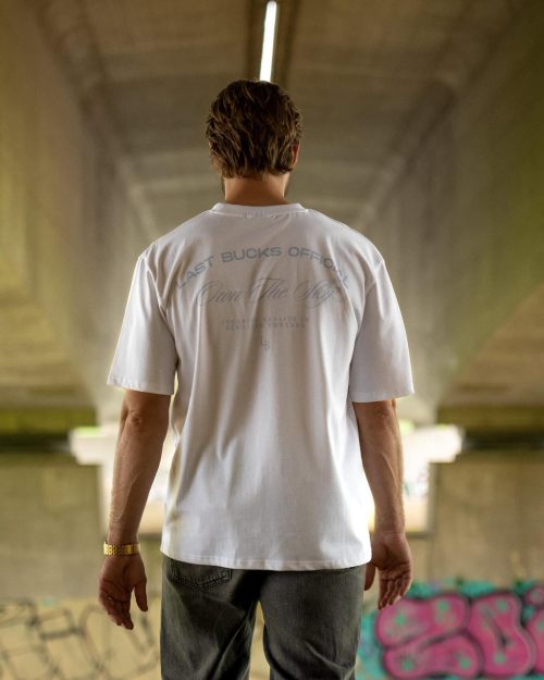 The founder wearing the Own The Sky Tee White by Last Bucks, back with the tonal Own The Sky script with Last Bucks Official, under a concrete overpass with graffiti