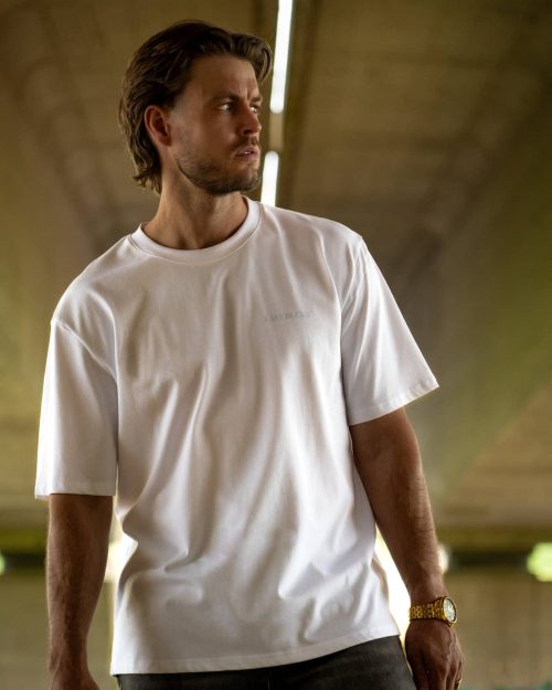 The founder wearing the Own The Sky Tee White by Last Bucks, front with the minimalist chest logo, under a concrete overpass with graffiti