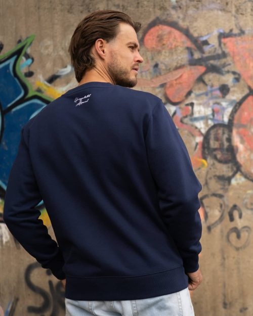 The founder wearing the Signature Sweater Navy by Last Bucks, back with the Unregulated Apparel embroidery at the back of the neck, against a concrete wall covered in graffiti