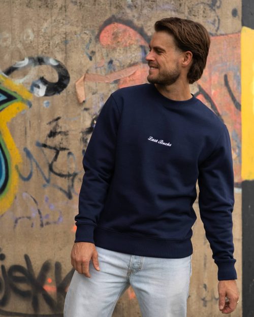 The founder wearing the Signature Sweater Navy by Last Bucks, front with the minimalist chest logo, against a concrete wall covered in graffiti
