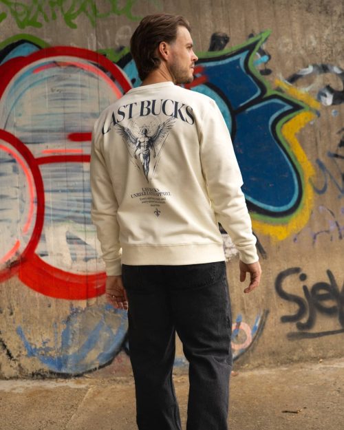 The founder wearing the Sinner & Saint Sweater Off-White by Last Bucks, back with a winged figure with Oscar Wilde's line, against a concrete wall covered in graffiti