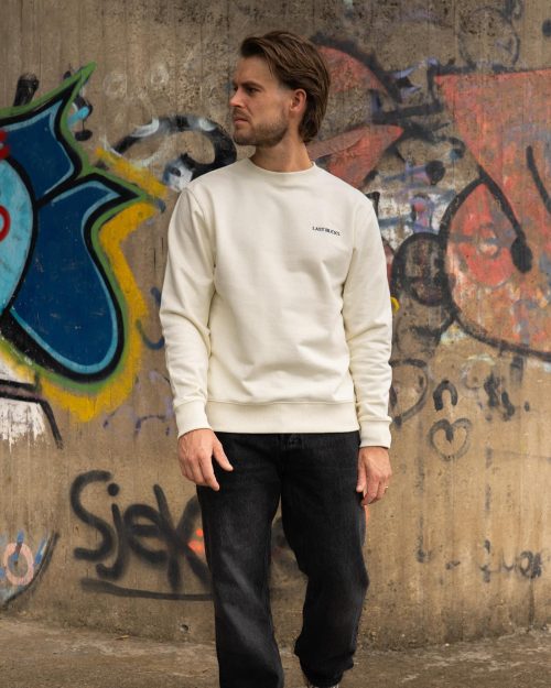 The founder wearing the Sinner & Saint Sweater Off-White by Last Bucks, front with the minimalist chest logo, against a concrete wall covered in graffiti