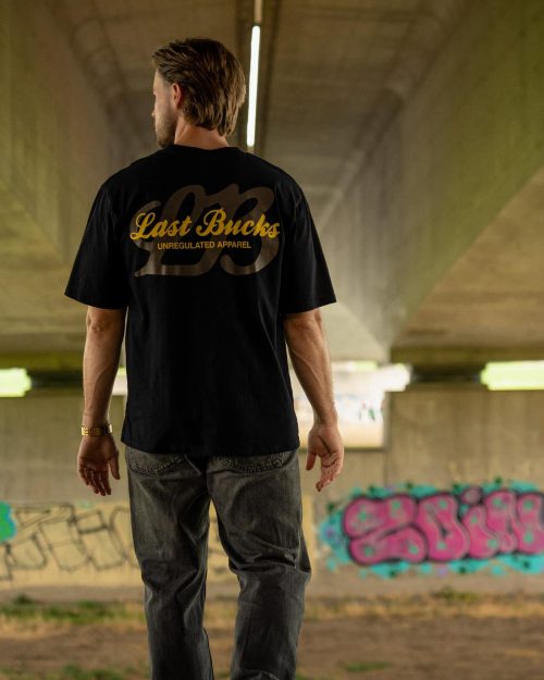 The founder wearing The Script Tee Jet Black by Last Bucks, back with Last Bucks in gold script with Unregulated Apparel over a tonal LB graphic, under a concrete overpass with graffiti