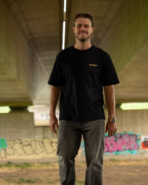 The founder wearing The Script Tee Jet Black by Last Bucks, front with the minimalist chest logo, under a concrete overpass with graffiti